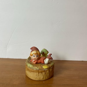 May include: A ceramic trinket box featuring a whimsical elf figure in a red outfit and a green mushroom. The box has a brown, textured exterior, and the elf is lying on a bed of straw. A charming decorative piece.