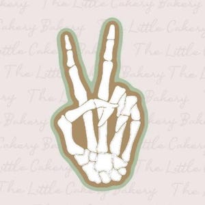 May include: A white skeleton hand making a peace sign, outlined in brown and light green. The background is a light pink with the words "The Little Cakery Bakery" in a cursive font.
