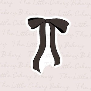 Puede incluir: Un cortador de galletas en forma de lazo negro con cintas largas y fluidas. El cortador tiene un contorno blanco y está sobre un fondo rosa claro con las palabras "The Little Cakery Bakery" en un patrón repetido.