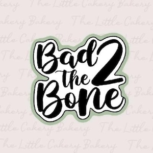 May include: A black and white cookie cutter with the text "Bad 2 the Bone" in a cursive font, outlined in black and a light green border. The background is a light pink with the words "The Little Cakery Bakery" repeated.