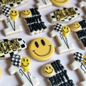 May include: A collection of decorated sugar cookies with various designs, including a smiley face, the words "One Happy Dude", and a checkered pattern. The cookies are white with black and yellow icing.