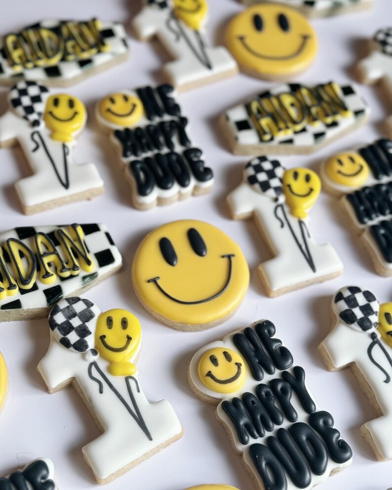 May include: A collection of decorated sugar cookies with various designs, including a smiley face, the words "One Happy Dude", and a checkered pattern. The cookies are white with black and yellow icing.
