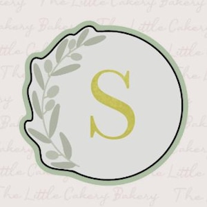 Peut inclure: Un emporte-pièce avec un bord vert clair et un intérieur gris clair. La lettre "S" est au centre en jaune-vert. Un motif de branche d'olivier vert clair est sur le côté gauche.