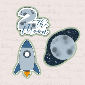 Puede incluir: Un conjunto de tres cortadores de galletas de temática espacial, de color azul y blanco. Los cortadores tienen forma de cohete, luna y el número 2 con las palabras "to the moon" escritas debajo.