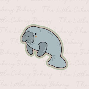 Puede incluir: Cortador de galletas de manatí azul claro con un diseño lindo. El manatí tiene un cuerpo gris, una cara sonriente y un contorno verde claro. El fondo tiene el texto "The Little Cakery Bakery".