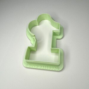 May include: A light green plastic cookie cutter shaped like the number one with the words "1 with balloons" on the bottom.