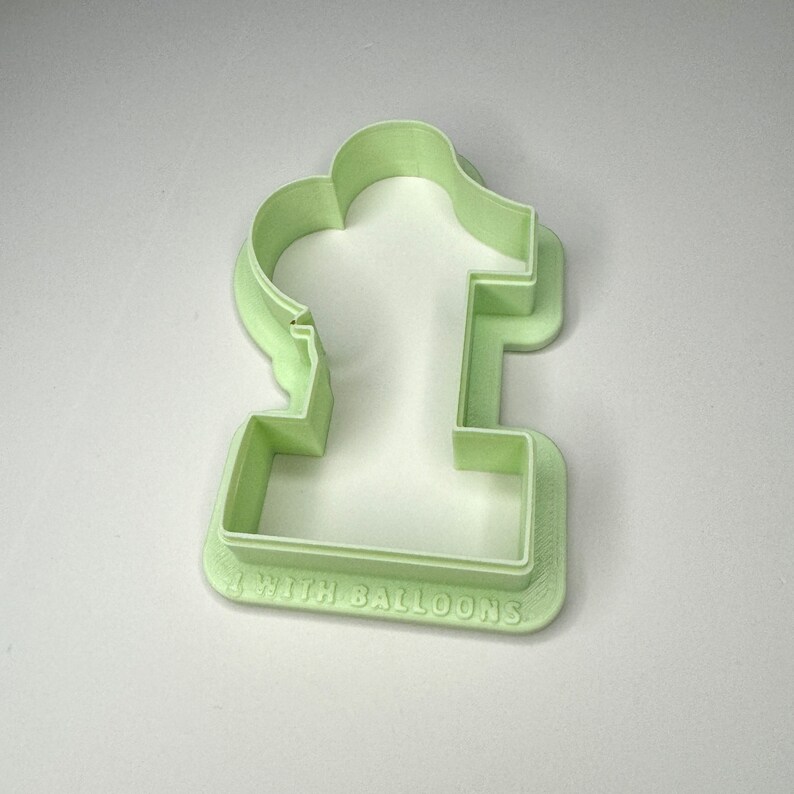May include: A light green plastic cookie cutter shaped like the number one with the words "1 with balloons" on the bottom.