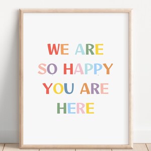 May include: A framed print with the words "WE ARE SO HAPPY YOU ARE HERE" in colorful block letters. The text is in red, orange, pink, blue, yellow, and green. The frame is a light wood color.