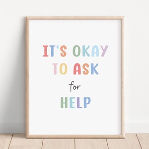 以下が含まれることがあります： 「IT'S OKAY TO ASK for HELP」という言葉がカラフルで遊び心のある文字で書かれた額入りのプリント。テキストは様々なパステルカラーで、白い背景に配置され、明るい木製のフレームに入っています。