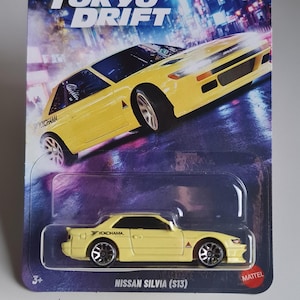 Puede incluir: Un coche de juguete fundido a presión Nissan Silvia (S13) amarillo de la serie Hot Wheels Silver, parte de la colección Fast & Furious: Tokyo Drift. El embalaje presenta el título de la película y un fondo de paisaje urbano.