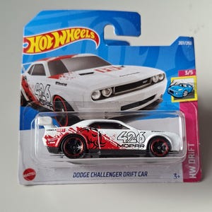 May include: A Hot Wheels Dodge Challenger Drift Car toy vehicle in white with red graphic accents and the number 426 printed on the side. The die-cast car is enclosed in its original blue and pink blister packaging with the text HW DRIFT.