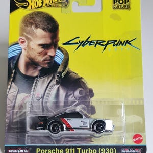 Może przedstawiać: Karta Hot Wheels Premium z odlewanym ciśnieniowo srebrnym samochodem Porsche 911 Turbo (930) z czerwonymi paskami i czarnymi kołami. Karta ma ż&oacute;łte tło z napisem "Cyberpunk 2077" i portretem mężczyzny. Karta ma r&oacute;wnież logo Hot Wheels i Mattel.