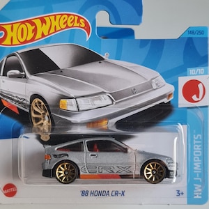 Puede incluir: Coche de juguete fundido a presión '88 Honda CR-X plateado con ruedas doradas, exhibido en una tarjeta Hot Wheels azul y blanca. El coche presenta gráficos CRX naranjas y negros. La tarjeta dice 'HW J-IMPORTS'.