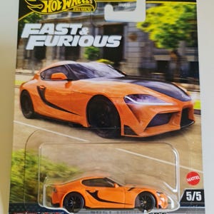 May include: A Hot Wheels die-cast model of an orange and black Toyota GR Supra from the Fast & Furious series. The car is in a clear plastic package with the Hot Wheels and Fast & Furious logos. The card reads "5/5".