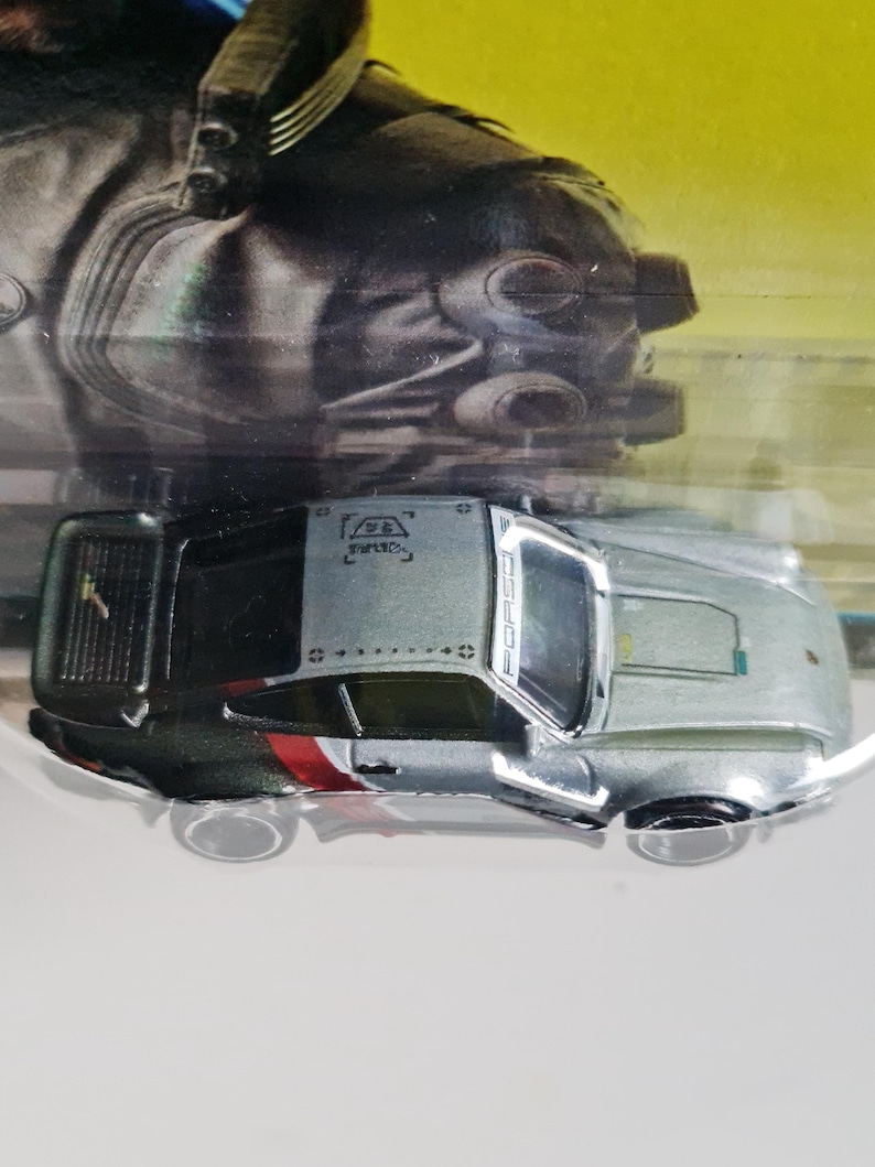 Może przedstawiać: Srebrno-czarny samochodzik z czerwono-białymi akcentami. Samoch&oacute;d ma napis "PORSCHE" z boku. Miniaturowa replika samochodu sportowego.