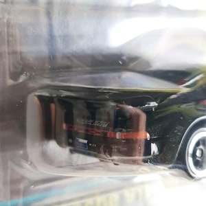 Może przedstawiać: Czarny samochodzik Porsche w przezroczystym plastikowym opakowaniu. Samoch&oacute;d ma czerwone i białe akcenty, widoczny jest napis "Porsche 911". Koła samochodu są srebrne.