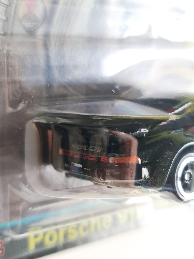 Może przedstawiać: Czarny samochodzik Porsche w przezroczystym plastikowym opakowaniu. Samoch&oacute;d ma czerwone i białe akcenty, widoczny jest napis "Porsche 911". Koła samochodu są srebrne.