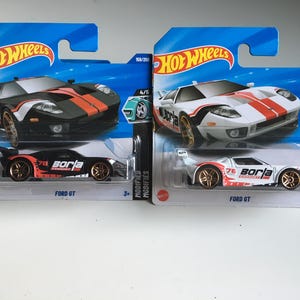 May include: Two Hot Wheels Ford GT model cars. One is black with orange stripes, the other is white with red stripes. Both have gold wheels and the "Borla" logo. The packaging is blue with the Hot Wheels logo.