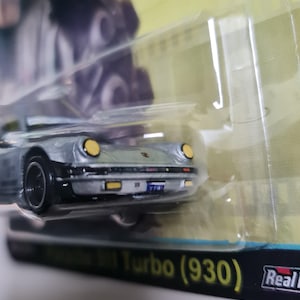 Może przedstawiać: Srebrny model samochodu Porsche 911 Turbo (930) z czarnymi kołami i ż&oacute;łtymi reflektorami. Samoch&oacute;d znajduje się w przezroczystym plastikowym opakowaniu z napisem "Real Riders" i "Turbo (930)".
