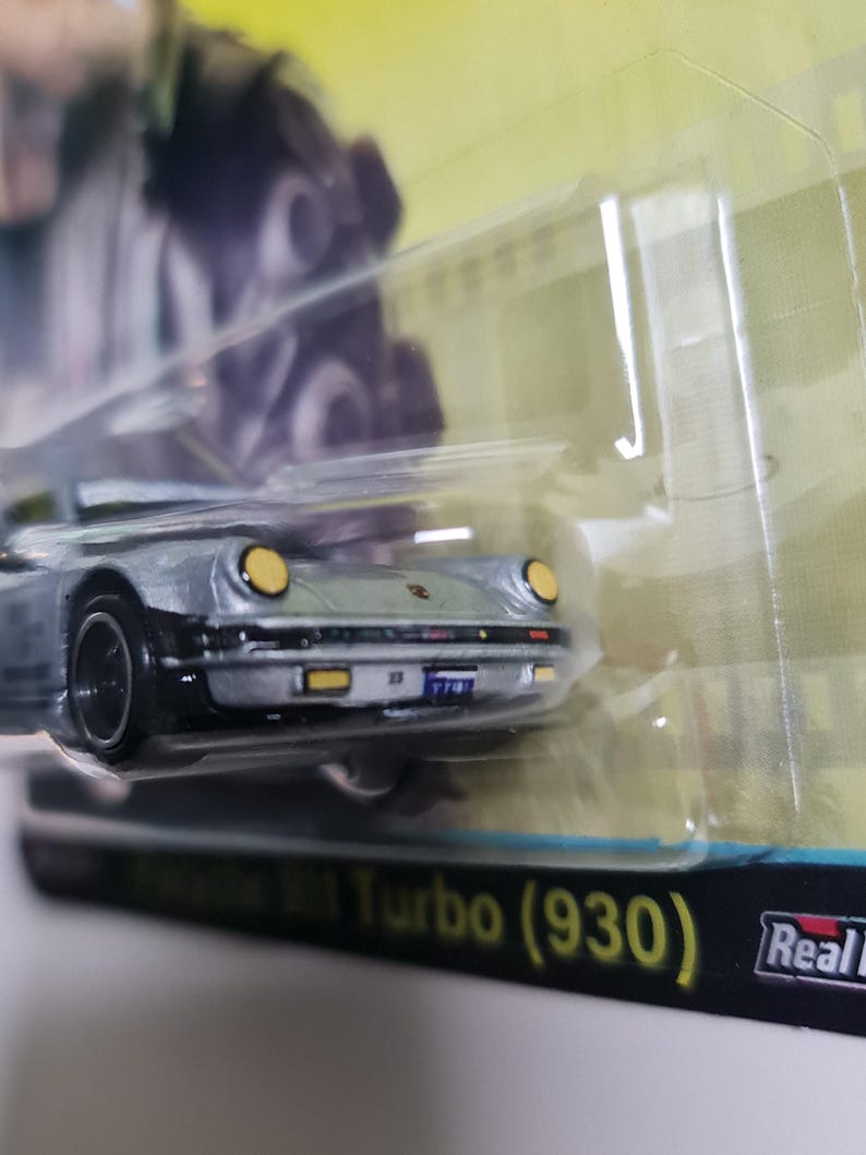 Może przedstawiać: Srebrny model samochodu Porsche 911 Turbo (930) z czarnymi kołami i ż&oacute;łtymi reflektorami. Samoch&oacute;d znajduje się w przezroczystym plastikowym opakowaniu z napisem "Real Riders" i "Turbo (930)".