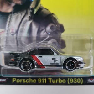 Może przedstawiać: Srebrny i czarny model odlewu Porsche 911 Turbo (930) z czerwono-białymi paskami wyścigowymi. Zabawkowe auto ma czarne koła i jest zapakowane w przezroczystą plastikową obudowę. Widoczny jest tekst "Porsche 911 Turbo (930)".