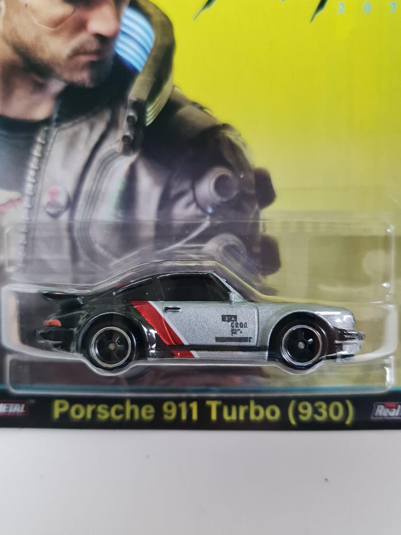 Może przedstawiać: Srebrny i czarny model odlewu Porsche 911 Turbo (930) z czerwono-białymi paskami wyścigowymi. Zabawkowe auto ma czarne koła i jest zapakowane w przezroczystą plastikową obudowę. Widoczny jest tekst "Porsche 911 Turbo (930)".