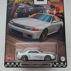 Może przedstawiać: Model Hot Wheels Premium Nissana Skyline GT-R (BNR32) w kolorze białym. Samochód zabawkowy jest prezentowany na karcie tematycznej z logo Hot Wheels i Boulevard. Karta przedstawia scenę z restauracji i numer 99.