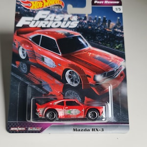 Może przedstawiać: Czerwony samochodzik Mazda RX-3 Hot Wheels z serii Fast & Furious. Samochód znajduje się na karcie z logo Fast & Furious i tłem miejskiego krajobrazu. Karta zawiera napis "Fast Rewind" i "1/5".