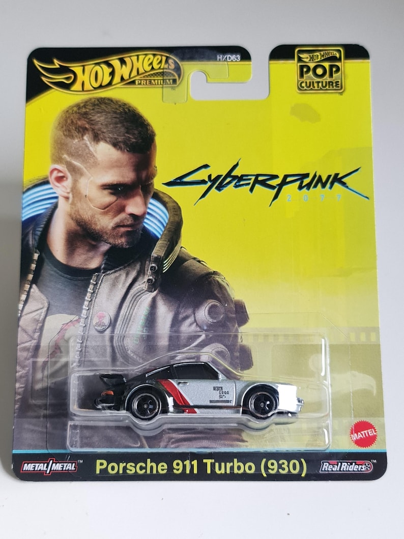Może przedstawiać: Karta Hot Wheels Premium z odlewanym ciśnieniowo srebrnym samochodem Porsche 911 Turbo (930) z czerwonymi paskami i czarnymi kołami. Karta ma ż&oacute;łte tło z napisem "Cyberpunk 2077" i portretem mężczyzny. Karta ma r&oacute;wnież logo Hot Wheels i Mattel.