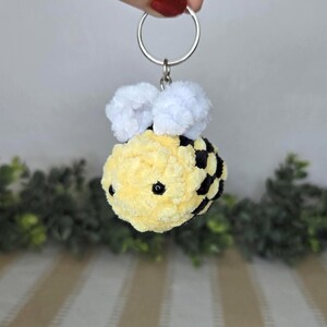 May include: A plush bee keychain featuring a soft, yellow and black striped body made of yarn. The bee has two small, white, textured wings on top and two tiny black bead eyes. It is attached to a circular silver metal split ring for keys.