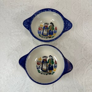 May include: Two ceramic bowls with a white interior and blue rim and handles. Each bowl has a colourful illustration of two people in traditional clothing. The bowls are set against a white lace background.