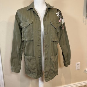 Puede incluir: Chaqueta utilitaria verde oliva con cierre de botones, múltiples bolsillos y bordado floral en el hombro. La chaqueta tiene un cuello clásico y mangas largas. La chaqueta se exhibe en un maniquí.