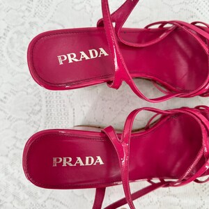 May include: A pair of bright pink Prada sandals. The sandals have a strappy design with thin straps and a high heel. The Prada logo is visible on the insole of each shoe. The shoes are made of a glossy material.