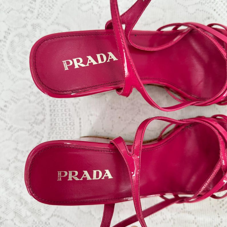May include: A pair of bright pink Prada sandals. The sandals have a strappy design with thin straps and a high heel. The Prada logo is visible on the insole of each shoe. The shoes are made of a glossy material.
