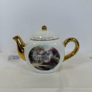 May include: White ceramic teapot with gold accents on the spout, handle, and lid knob. The teapot features a scenic image of a house and the text "HOME IS WHERE THE HEART IS".