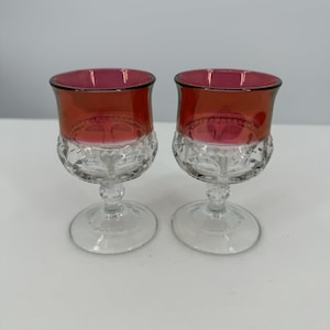 Puede incluir: Dos copas de vino vintage con cuencos de color rojo rubí y bases transparentes estampadas. Las copas tienen un diseño clásico con un cuenco redondeado y una base con pie. El color rojo rubí añade un toque de elegancia a la cristalería.