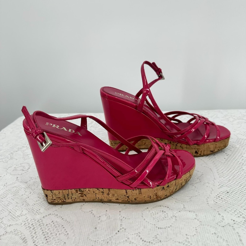 May include: Pair of bright pink Prada wedge sandals. The sandals feature a glossy finish, multiple straps, and cork platforms. The Prada logo is visible on the side of the sandal. The wedge heel measures approximately 4 inches.