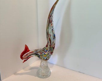 Könnte beinhalten: Eine farbenfrohe Glasskulptur eines Vogels mit langem, gebogenem Hals und Schwanz. Der Vogel hat einen roten Kopf und Schwanz, der Körper ist mit verschiedenen Farben gesprenkelt. Er steht auf einem klaren, gedrehten Glassockel. Die Skulptur ist etwa 30 cm hoch.