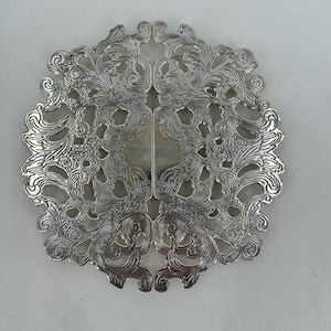 Puede incluir: Salvamanteles de metal plateado ornamentado con un diseño calado simétrico. El salvamanteles presenta patrones florales y de volutas, creando una apariencia decorativa y elegante. El diseño está centrado con una sección rectangular.