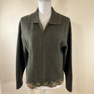 May include: An olive green zip-up jacket with a collar and long sleeves. The jacket features decorative stitching around the collar and a detailed embroidered design of pinecones and greenery along the bottom hem. The jacket is displayed on a mannequin.