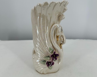 May include: A pearlescent white swan vase with a ruffled top. The vase features a swan design with gold accents and purple flower and green leaf details. A decorative piece for home decor.