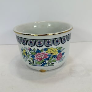 Op de afbeelding: Witte keramische plantenbak met een gouden rand en voet. De plantenbak heeft een bloemenontwerp met roze, gele en blauwe bloemen. Een decoratieve band met een blauwe achtergrond en bloemmotief bevindt zich bij de rand.