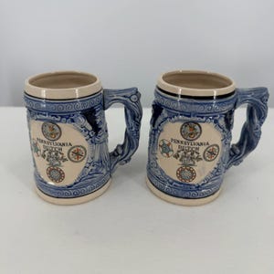 May include: Two ceramic beer mugs featuring a cream-colored body with blue relief detailing. Each mug displays the text Pennsylvania Dutch, surrounded by circular motifs and a central illustration of two figures, finished with a decorative handle.
