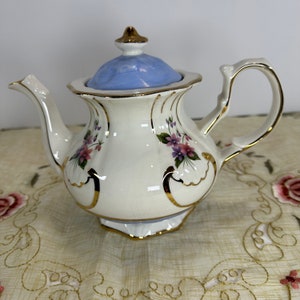 May include: A vintage porcelain teapot with a light blue lid and gold trim. The teapot is decorated with purple and pink floral designs. The handle and spout are also trimmed in gold. The teapot is sitting on a floral patterned tablecloth.