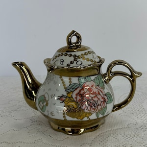 Puede incluir: Una pequeña tetera decorativa con boquilla, asa y adornos dorados. El cuerpo blanco presenta un diseño floral con rosas rosas y amarillas, acentuado con detalles dorados. La tapa también es dorada con un adorno decorativo.