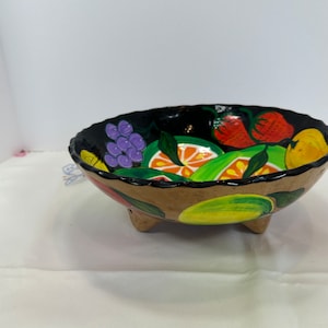 May include: A decorative fruit bowl with a black interior and a tan exterior. The bowl is adorned with painted images of various fruits, including grapes, strawberries, and citrus slices. The bowl has three small feet.