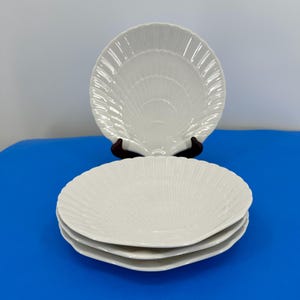 Puede incluir: Cuatro platos blancos en forma de concha apilados sobre una superficie azul. Los platos tienen un diseño texturizado en forma de abanico. Un plato se muestra en un soporte marrón oscuro. Los platos son de cerámica.