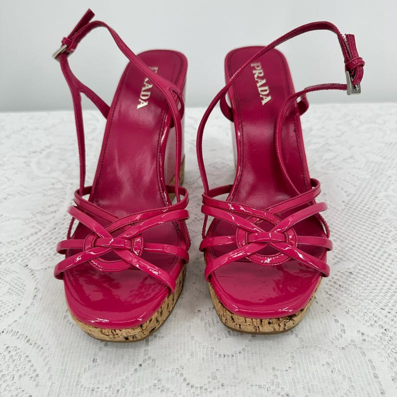 May include: Pair of bright pink Prada wedge sandals. The sandals feature a glossy finish, multiple straps, and a cork wedge heel. The ankle straps have silver buckles. The Prada logo is visible on the side of the heel.