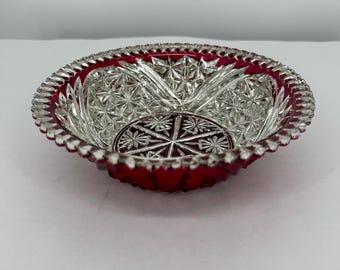 May include: A clear glass bowl with a ruby red rim and base. The bowl has a detailed cut glass design with star and geometric patterns. The rim has a scalloped edge. The bowl is likely vintage, and would be measured in centimetres.