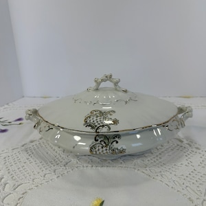 Może przedstawiać: Biała porcelanowa salaterka z pokrywką. Naczynie ma złote obramowanie i dekoracyjne złote i czarne wzory kwiatowe. Pokrywka ma ozdobny uchwyt. Naczynie ma owalny kształt z uchwytami po bokach.